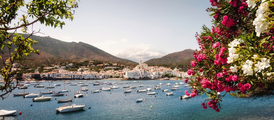 Küstenstadt mit Booten im Hafen, umgeben von Blumen und Bergen.
